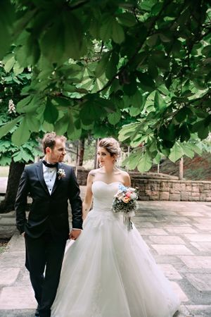 A couple walks together under green leafy trees in wedding attire on stone pavement.