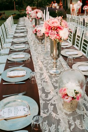 Elegant wedding table setting with pink floral centerpieces and lace runner on dark wooden table.