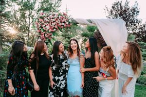 Women in festive dresses gathered around floral arch at outdoor celebration.