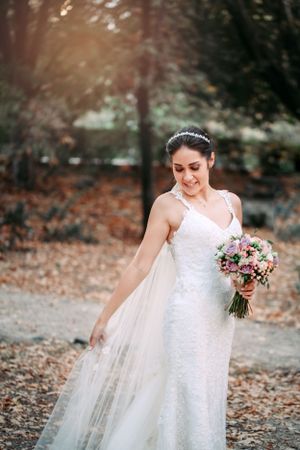 Bride in a lace wedding dress holding a pink bouquet while standing on fallen autumn leaves.