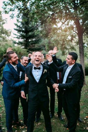 Groomsmen in dark suits celebrating and laughing together outdoors.