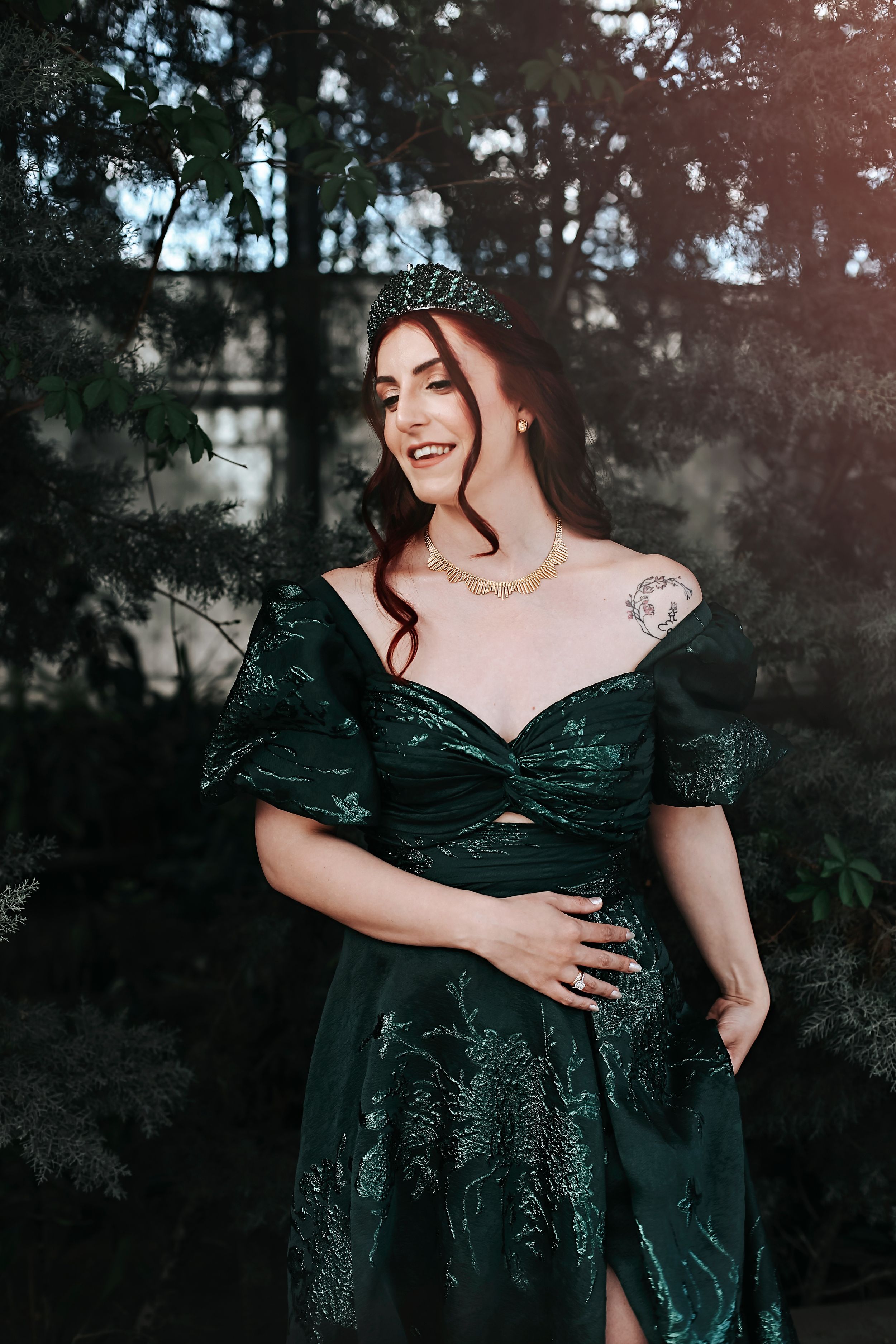 A woman in a dark green gown poses outdoors in a moody forest setting while wearing a tiara.