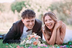 Couple lying on grass laughing together with wedding bouquet.