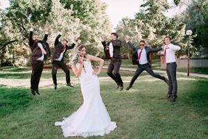 Fun wedding party photo with groomsmen jumping in background while bride poses.
