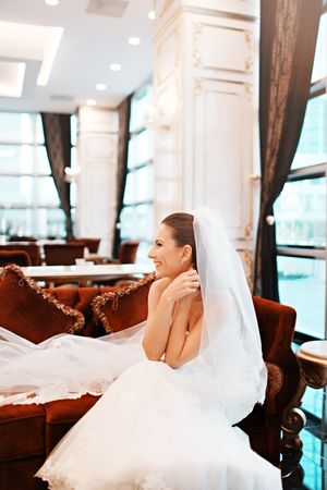 A bride in a white gown relaxes on an elegant brown leather couch in a bright venue.