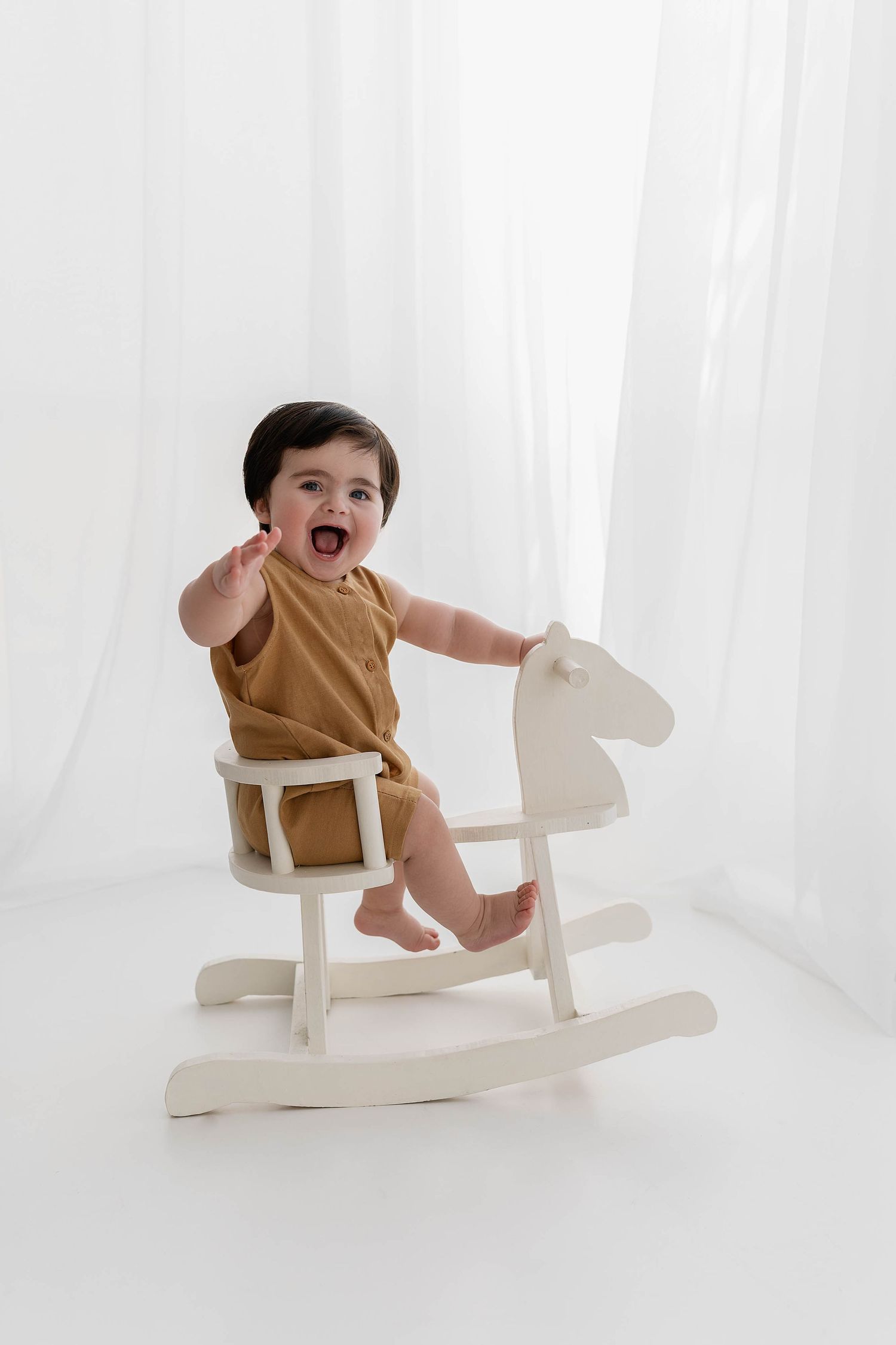 Excited baby in brown romper sitting on white wooden rocking horse.