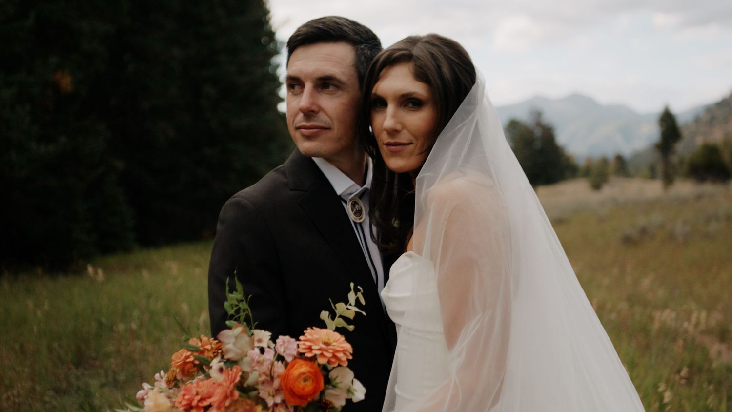 A couple in wedding attire embraces in a scenic mountain meadow with a cloudy sky backdrop.