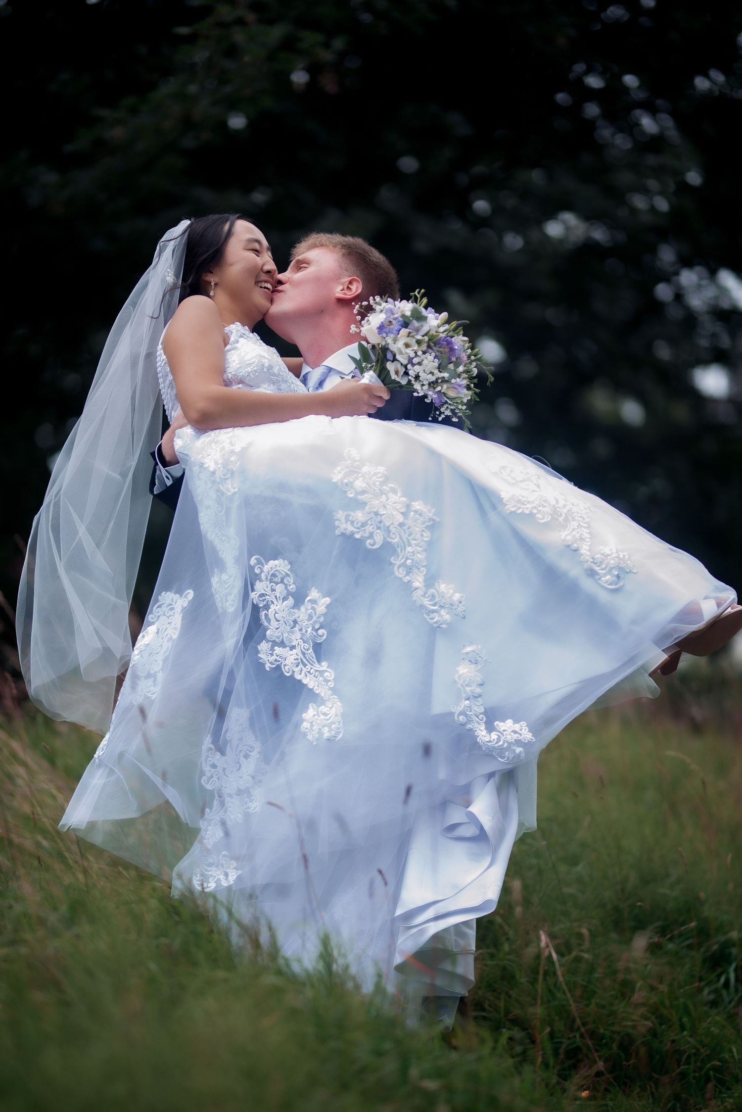 groom carries bride in his arms kissing her on the cheeks as she smiles holding boquet of purple and white flowers