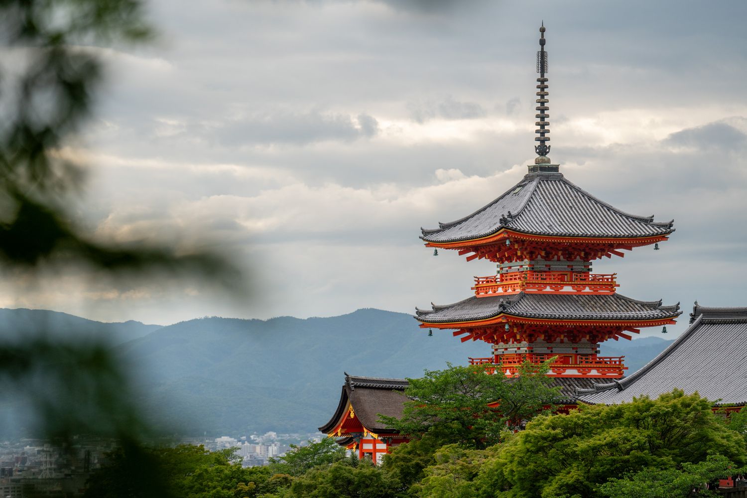 Red pagoda tower of Kiyomizu-dera temple emerging from forest against misty mountain backdrop.