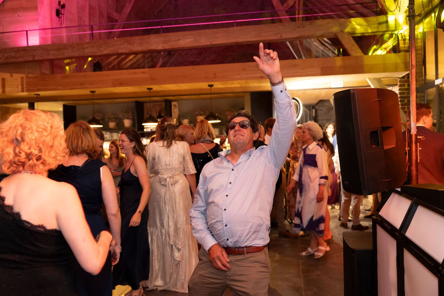 Wedding guests dancing energetically at a reception in a rustic venue with warm lighting.