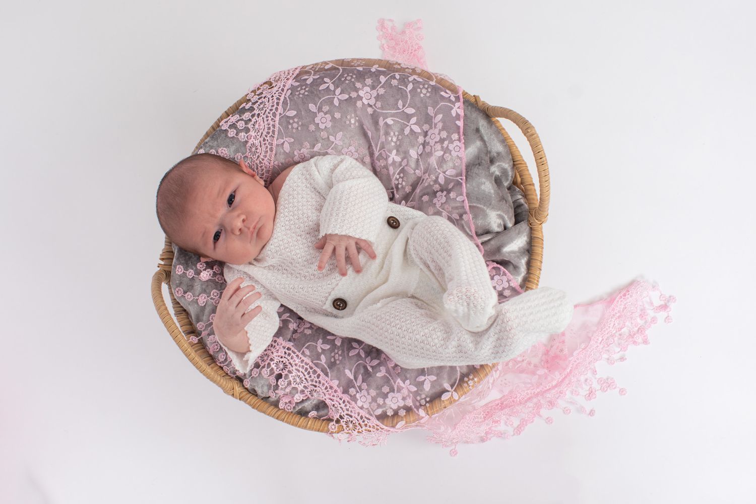 A newborn baby rests in a woven basket with pink and grey textured bedding.