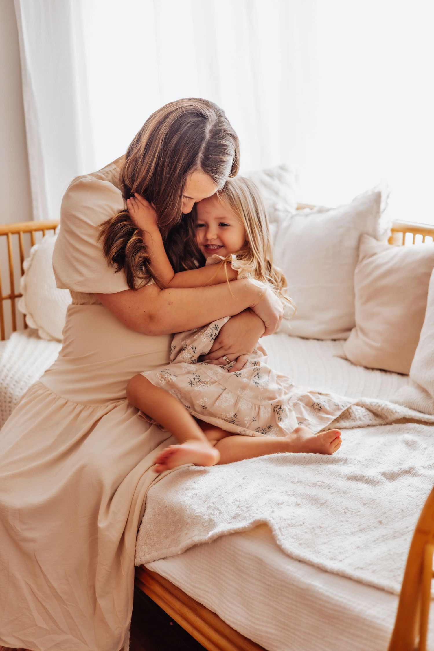A tender moment of cuddling and laughter on a bright white bed with natural lighting streaming through curtains. Family sessions