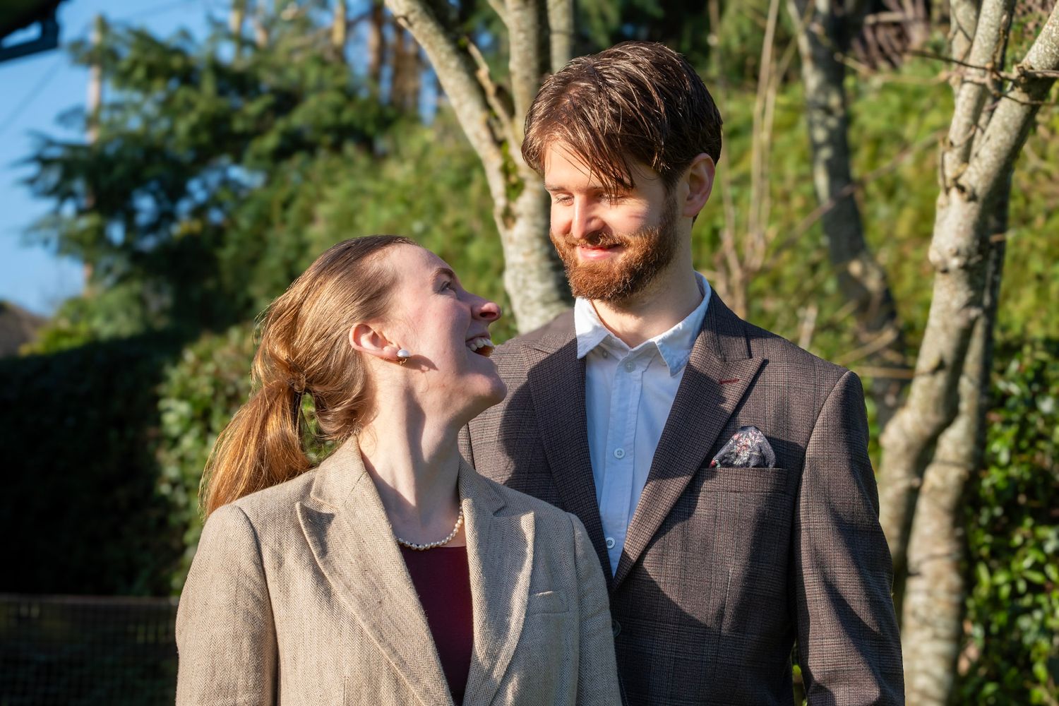 A couple in formal attire share a sweet moment together in a sunny garden setting with trees in the background.