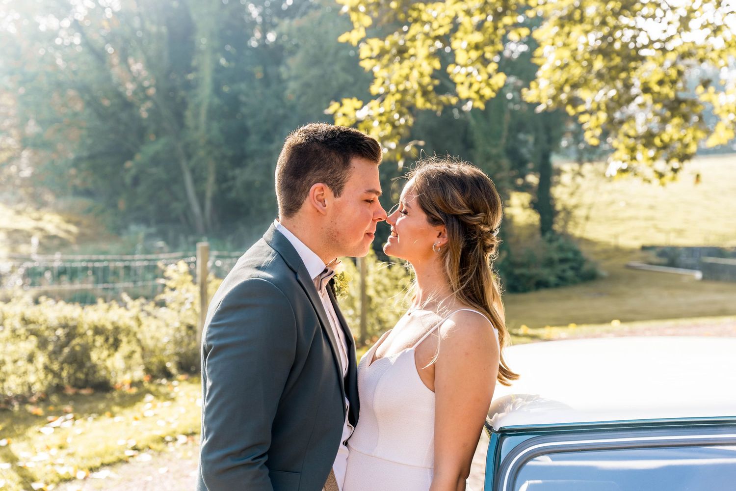 A romantic sunlit portrait beside a classic car during an outdoor wedding shoot.
