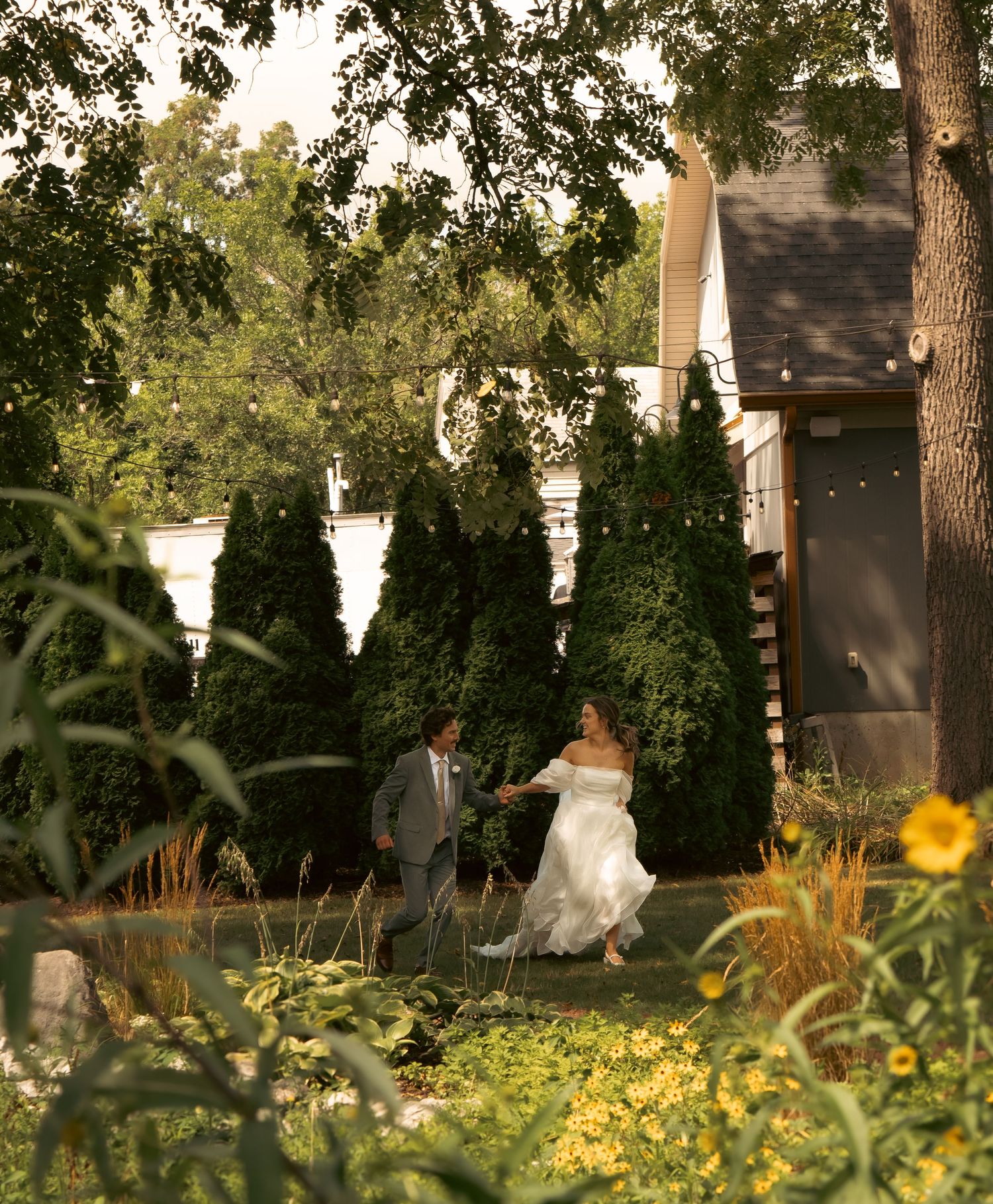 Newlyweds walk through a garden path with string lights and evergreen trees creating a romantic atmosphere.