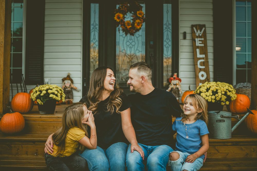 A fall front porch scene decorated with pumpkins, mums and a sunflower wreath as a family sits on the steps. Bon Accord Family Photographer