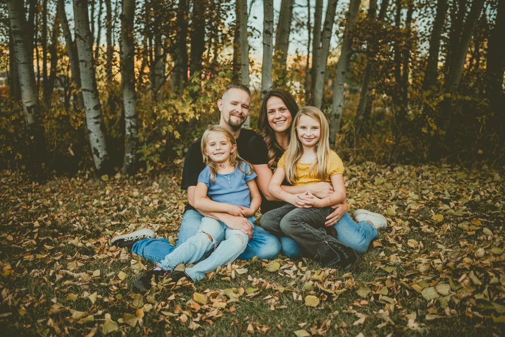 A group sits together on fallen autumn leaves in a wooded area during golden hour. Bon Accord Family Photographer