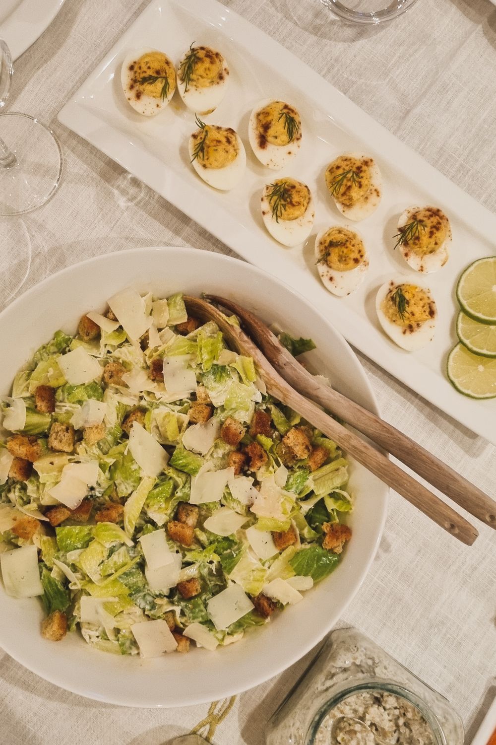 Caesar salad and deviled eggs garnished with fresh herbs and lime wedges on white serving plates.
