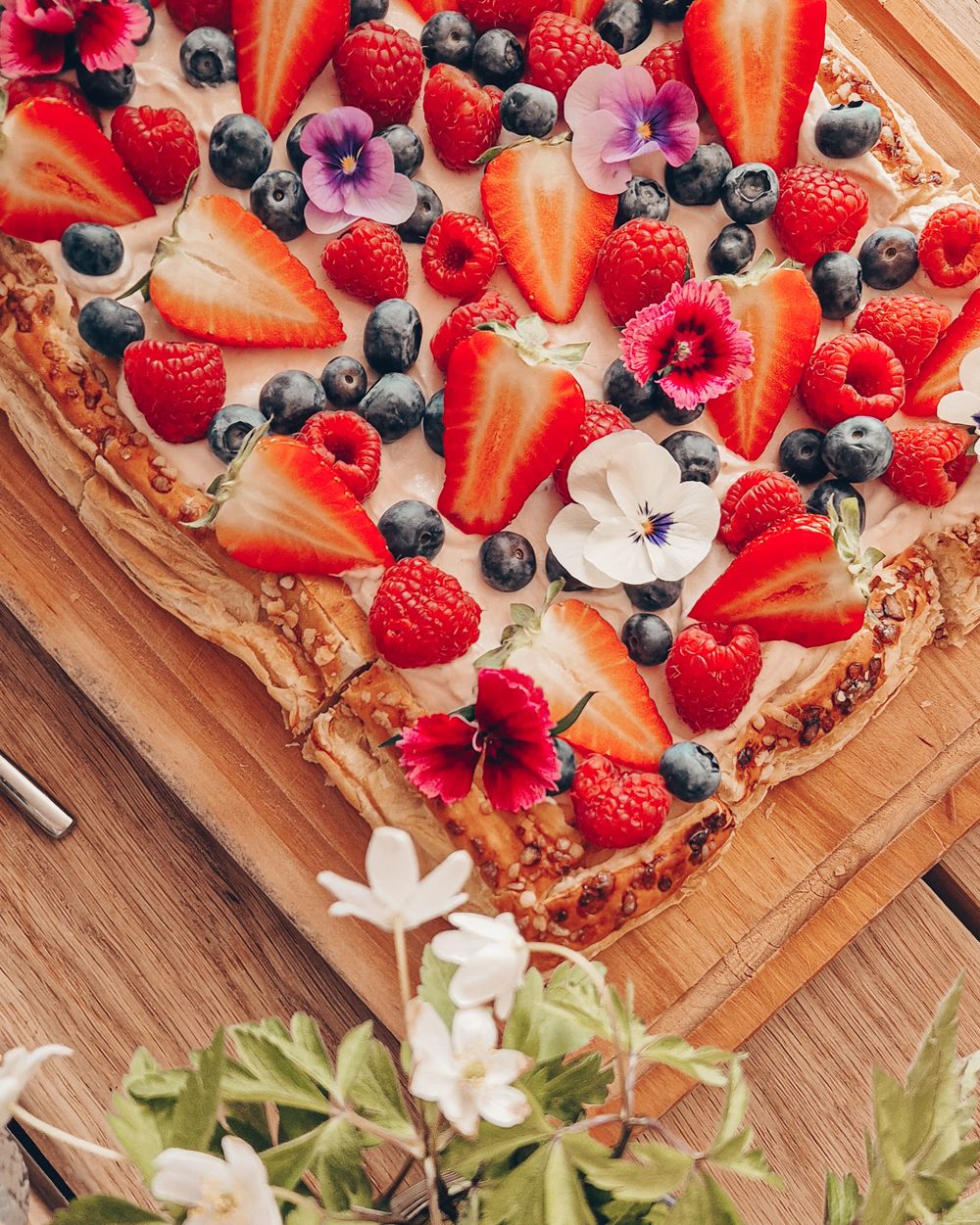 Fresh berry tart topped with strawberries, blueberries, raspberries and edible flowers on a wooden cutting board.