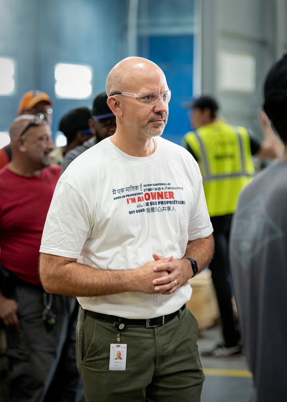Professional in a white t-shirt and khaki pants stands in an industrial or warehouse setting.