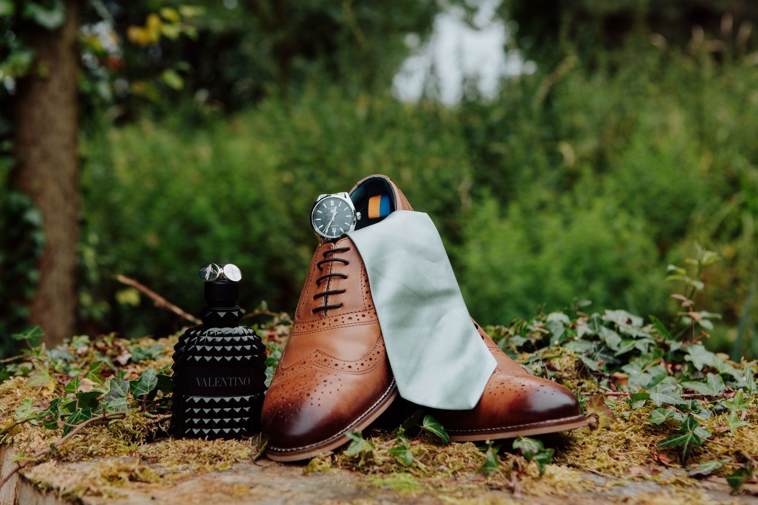 Elegant brown leather dress shoe and accessories arranged on forest floor.