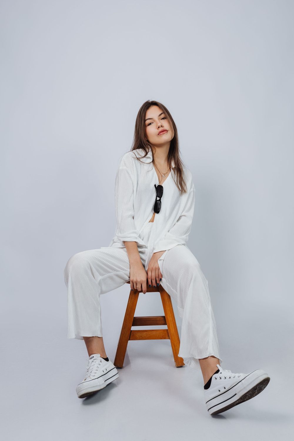 Model in all-white outfit and sneakers poses casually on wooden stool against grey background.