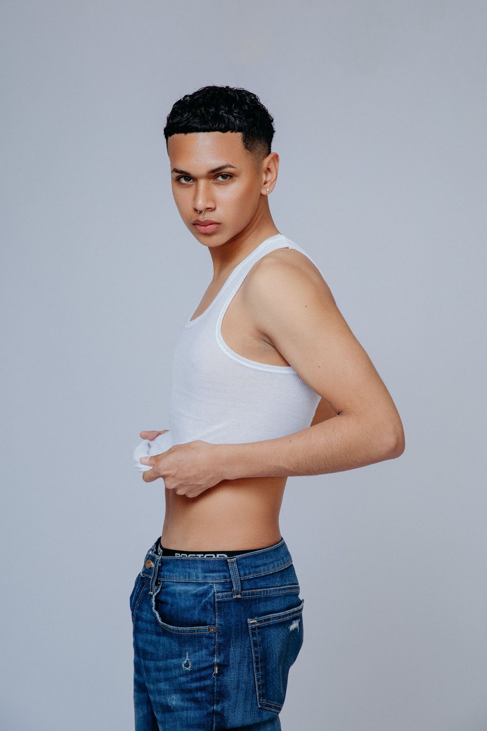 Athletic pose in white tank top and jeans against grey studio background.
