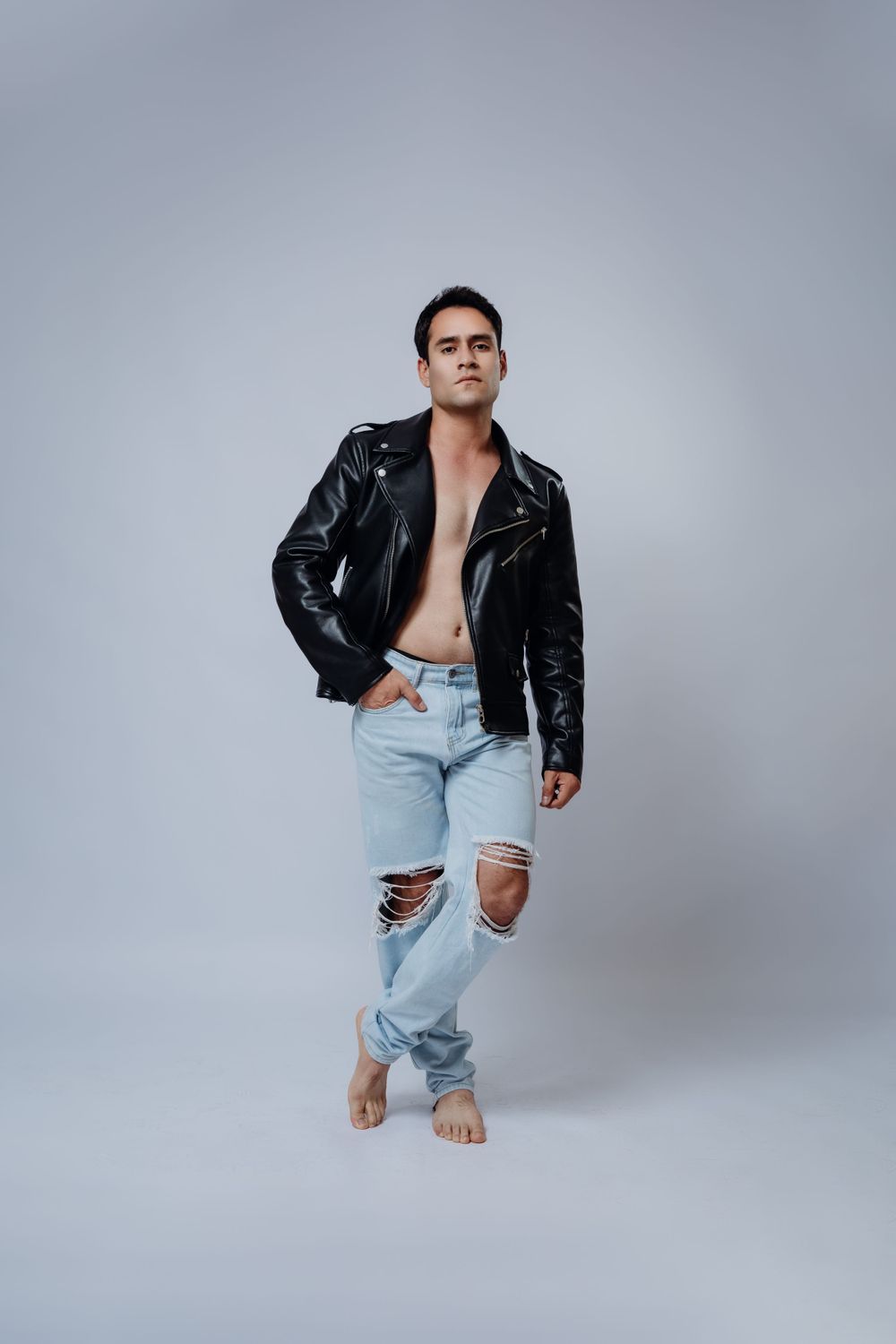 Fashion model in leather jacket and distressed jeans against grey studio background.