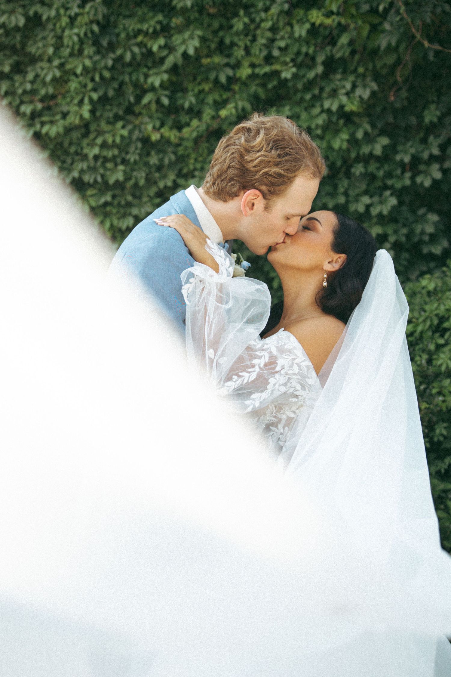 Romantic wedding portrait through flowing veil against ivy-covered wall.