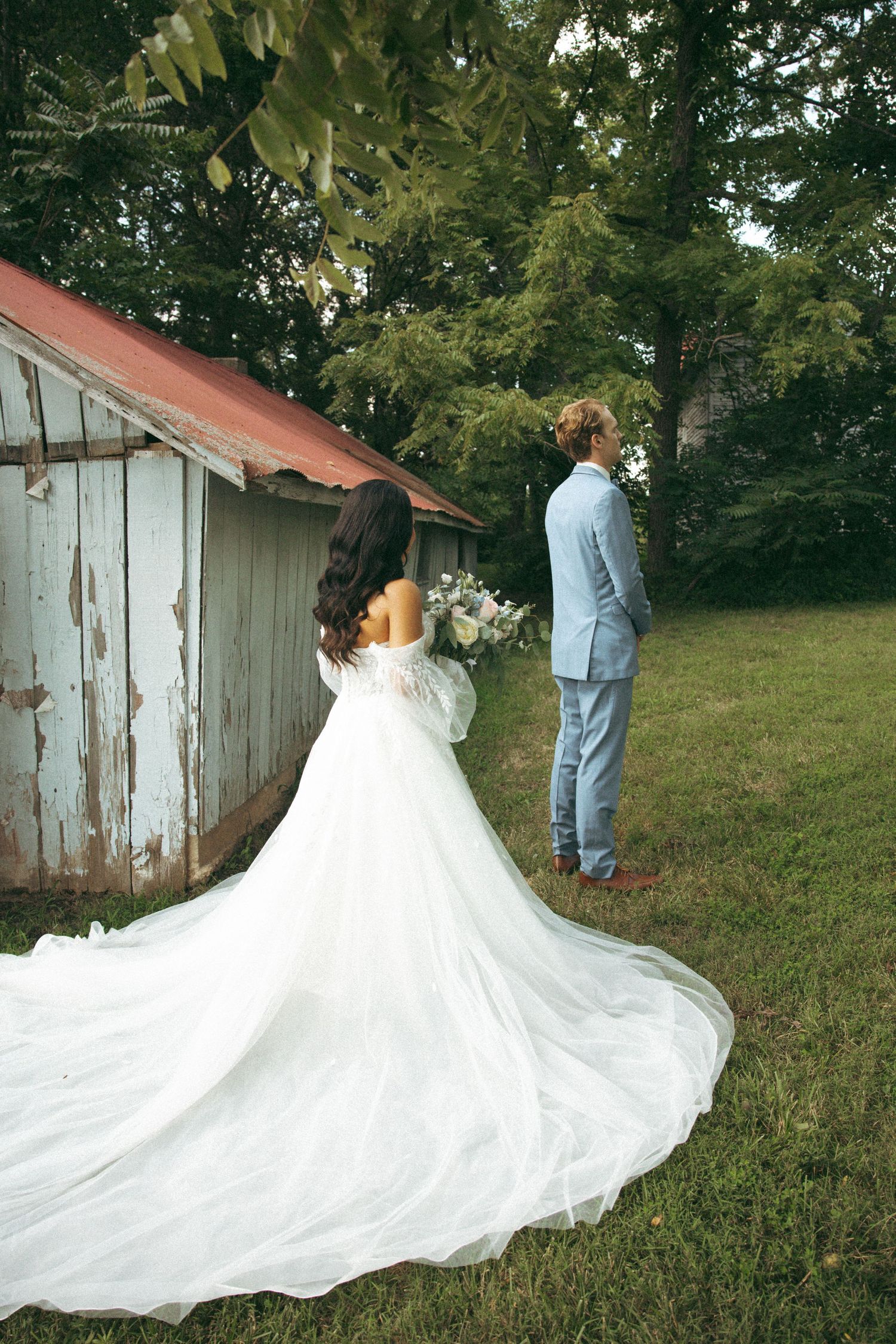 Wedding dress with dramatic train trails behind couple standing near rustic barn.