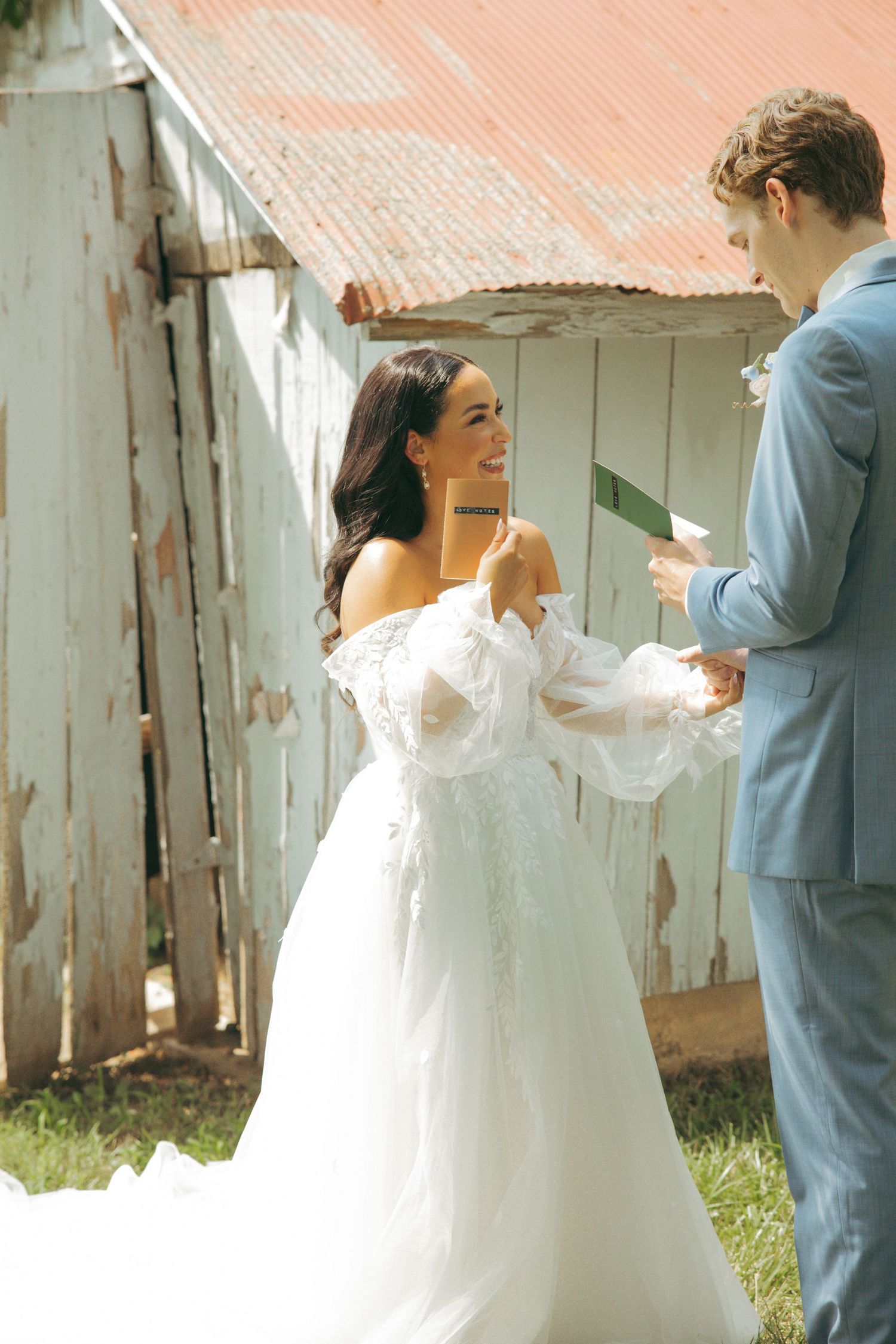 Wedding couple exchanges vows or notes beside weathered white barn wall.