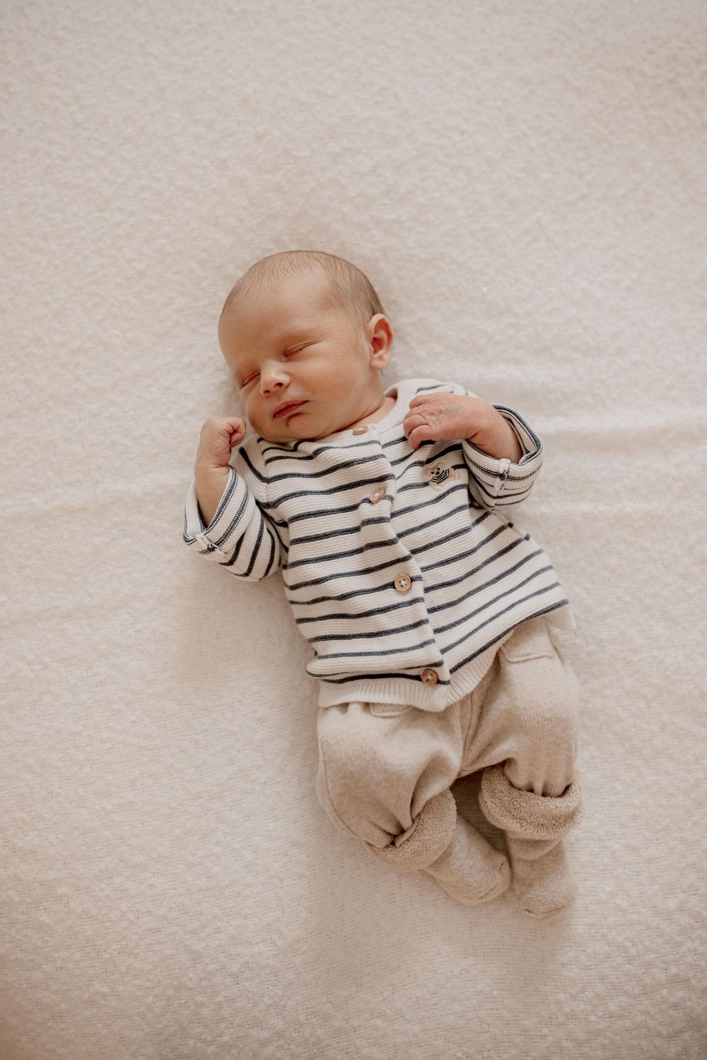 Sleeping baby wearing striped shirt and beige pants lying on white blanket in peaceful slumber.