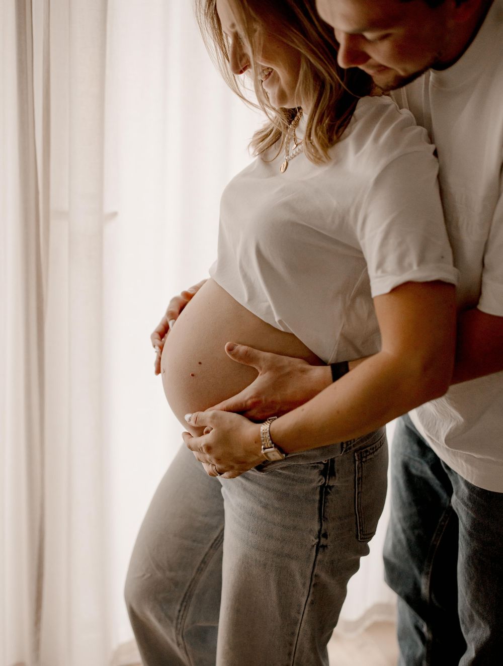 Couple shares intimate moment near window as they embrace a pregnant belly in casual white shirts and jeans.