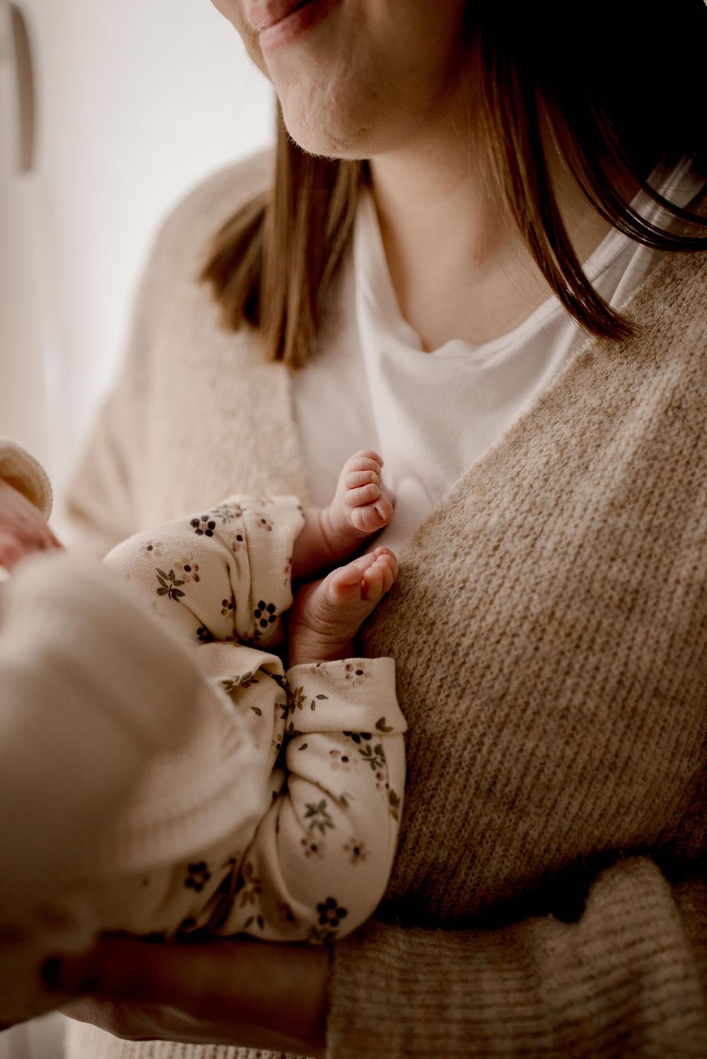 A tiny baby's hand grasps while wearing a floral onesie against someone in a cozy beige sweater.