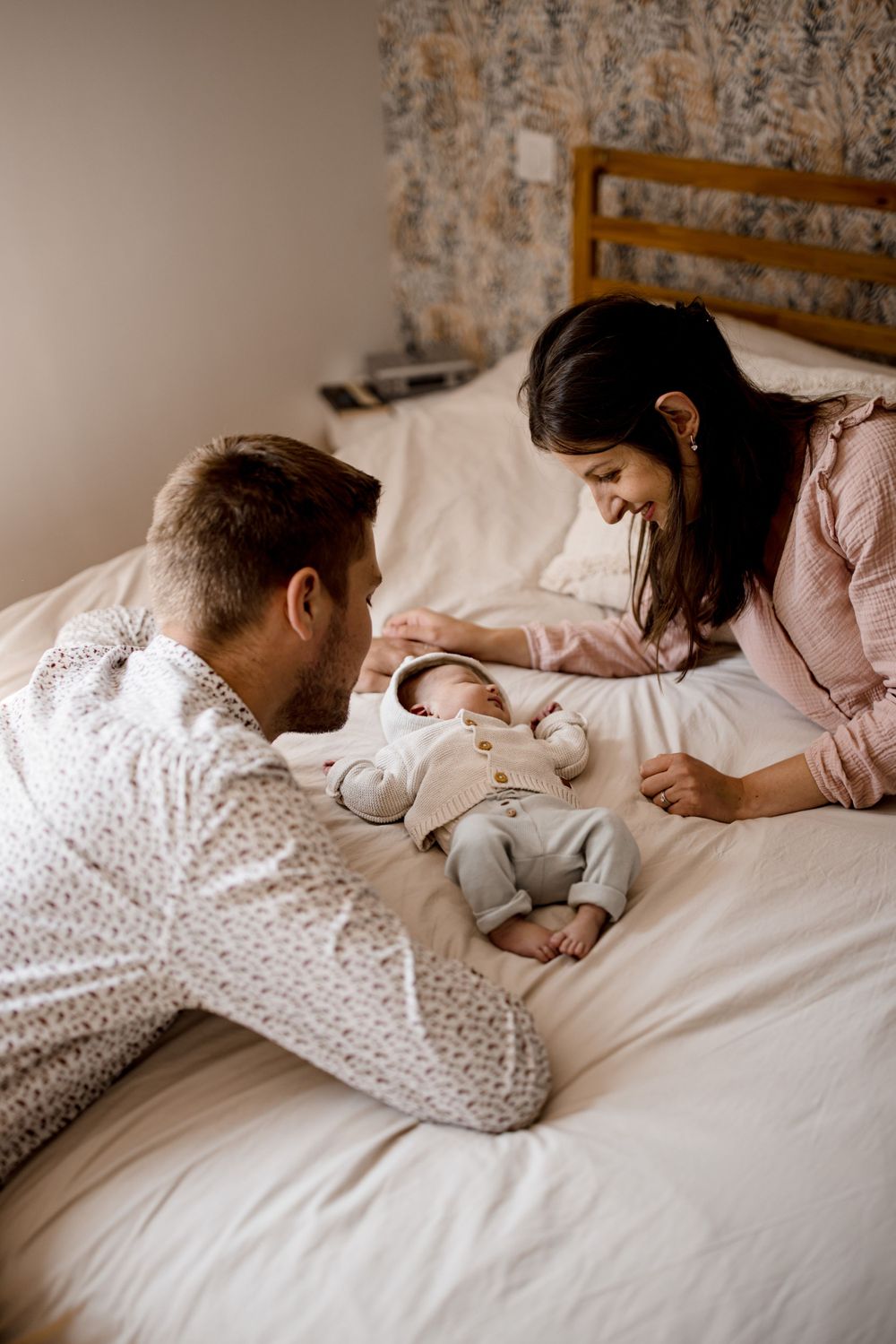 Un moment de tendresse en famille sur un lit avec du papier peint fleuri en arrière-plan.