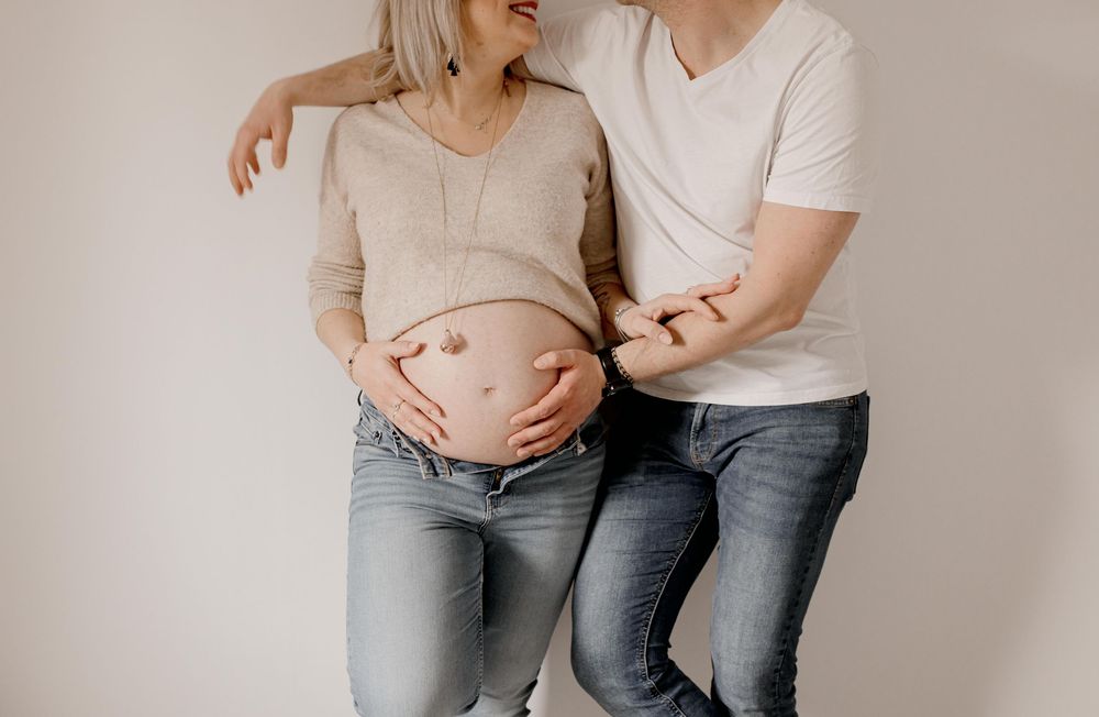 Couple in casual clothing tenderly embraces while holding pregnant belly against neutral background.