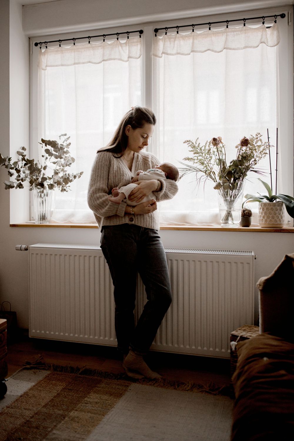 A figure stands by a window with sheer curtains holding a baby, surrounded by potted plants in a cozy room setting.