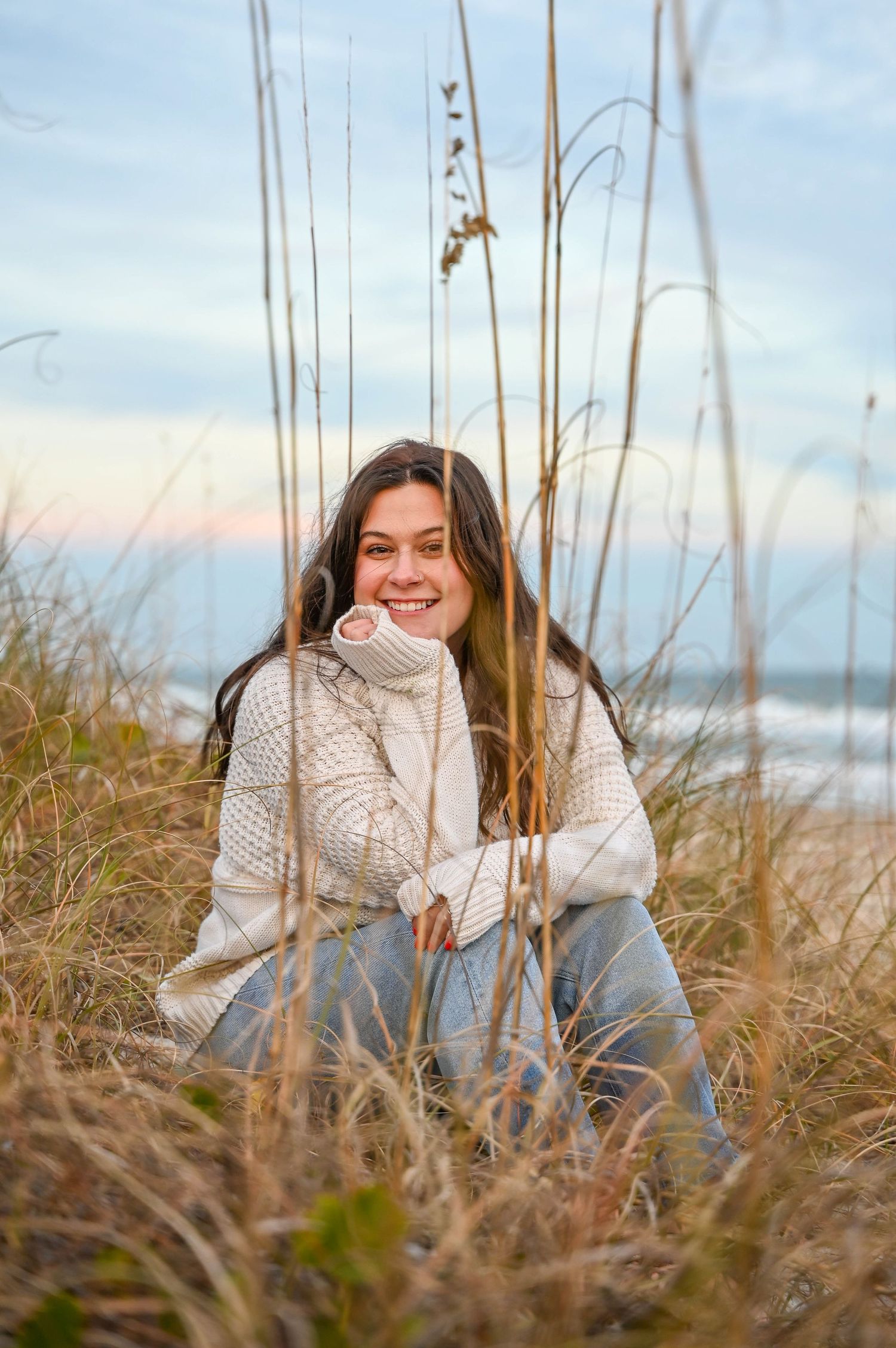Young woman in cozy sweater sitting among beach grass during sunset.