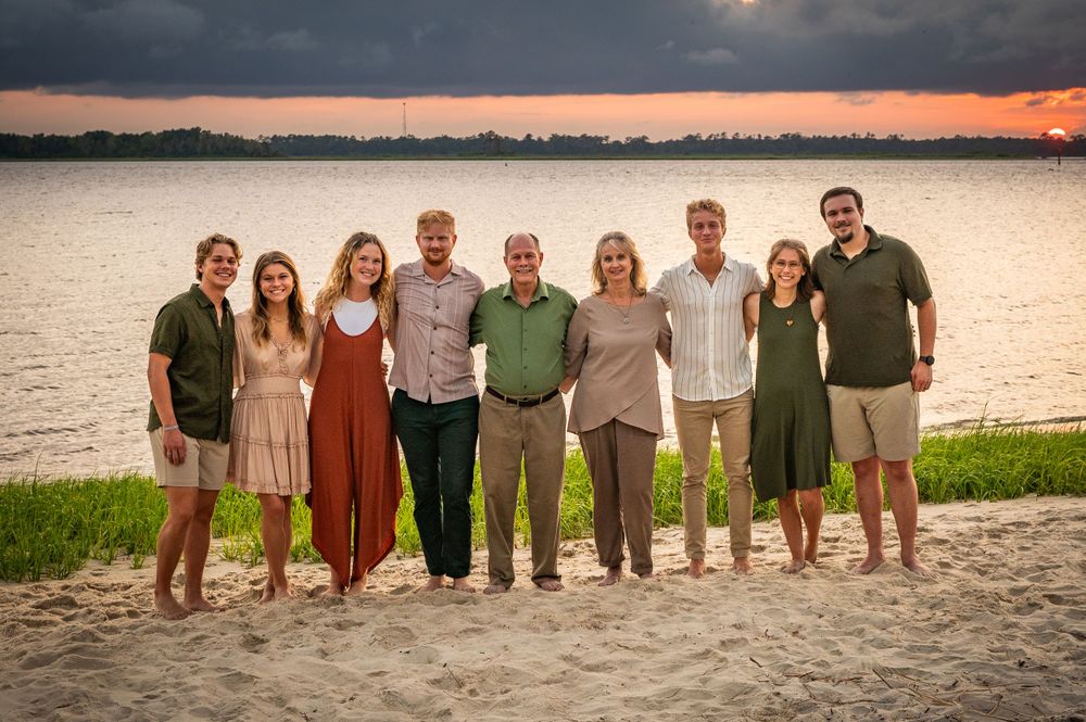 Family group portrait on beach at sunset with water and dramatic sky in background.
