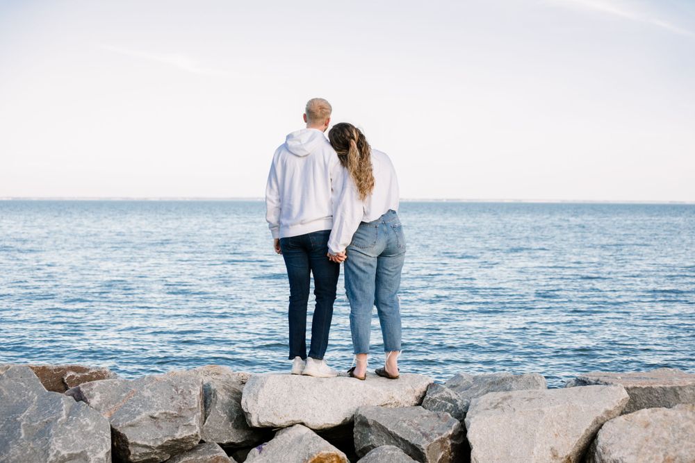 Couple standing together on rocky shoreline overlooking calm ocean waters.