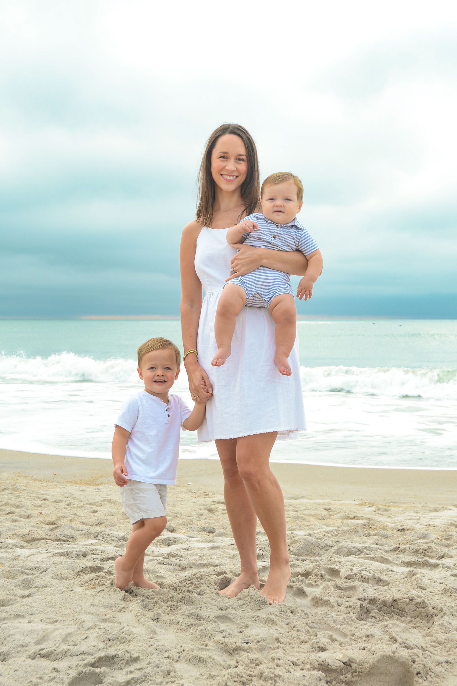 A family in white attire poses together on a sandy beach with ocean waves in the background.