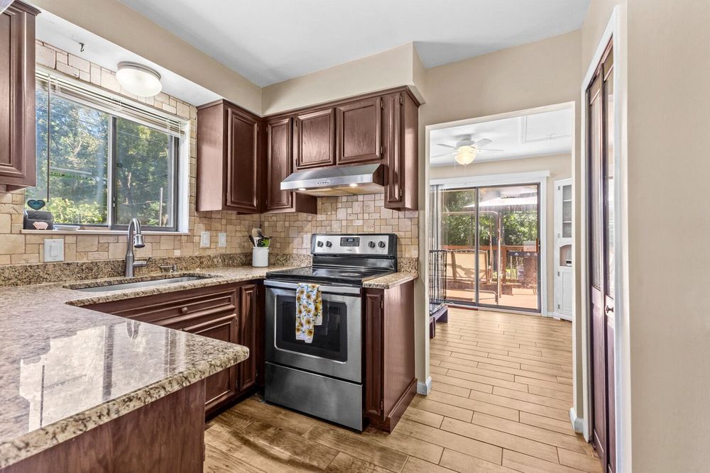 Modern kitchen with dark wood cabinets, granite countertops, stainless steel range, and tile backsplash leading to a sunlit patio.