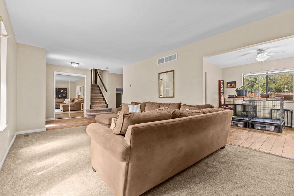 Spacious living room with beige carpet, neutral walls, brown sofa, ceiling fan, and staircase visible in background.