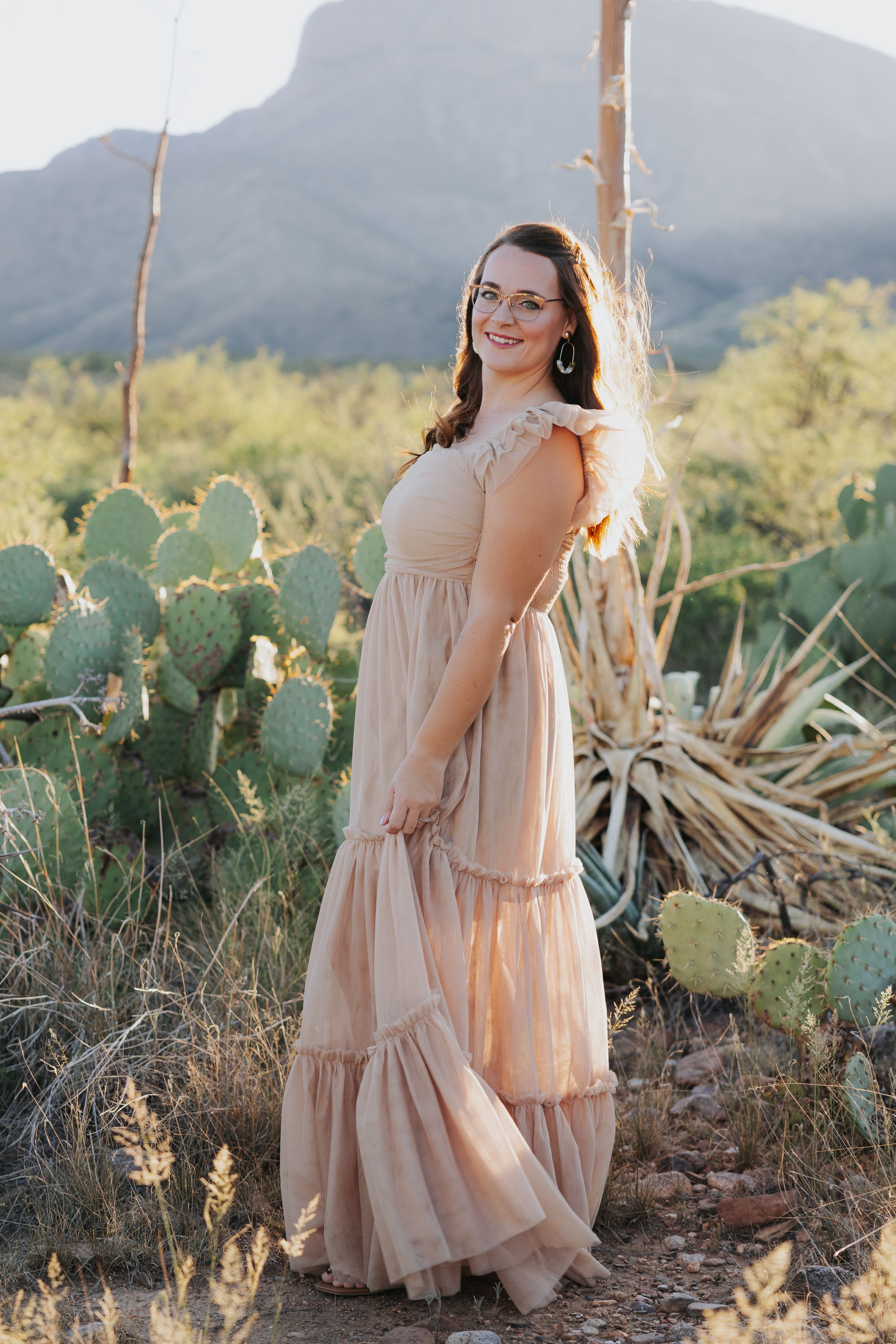 Desert photoshoot featuring a flowy beige dress among prickly pear cactus and mountains.