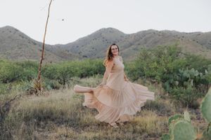 A flowing peach dress twirls gracefully against a desert mountain backdrop with cacti and dry grass.