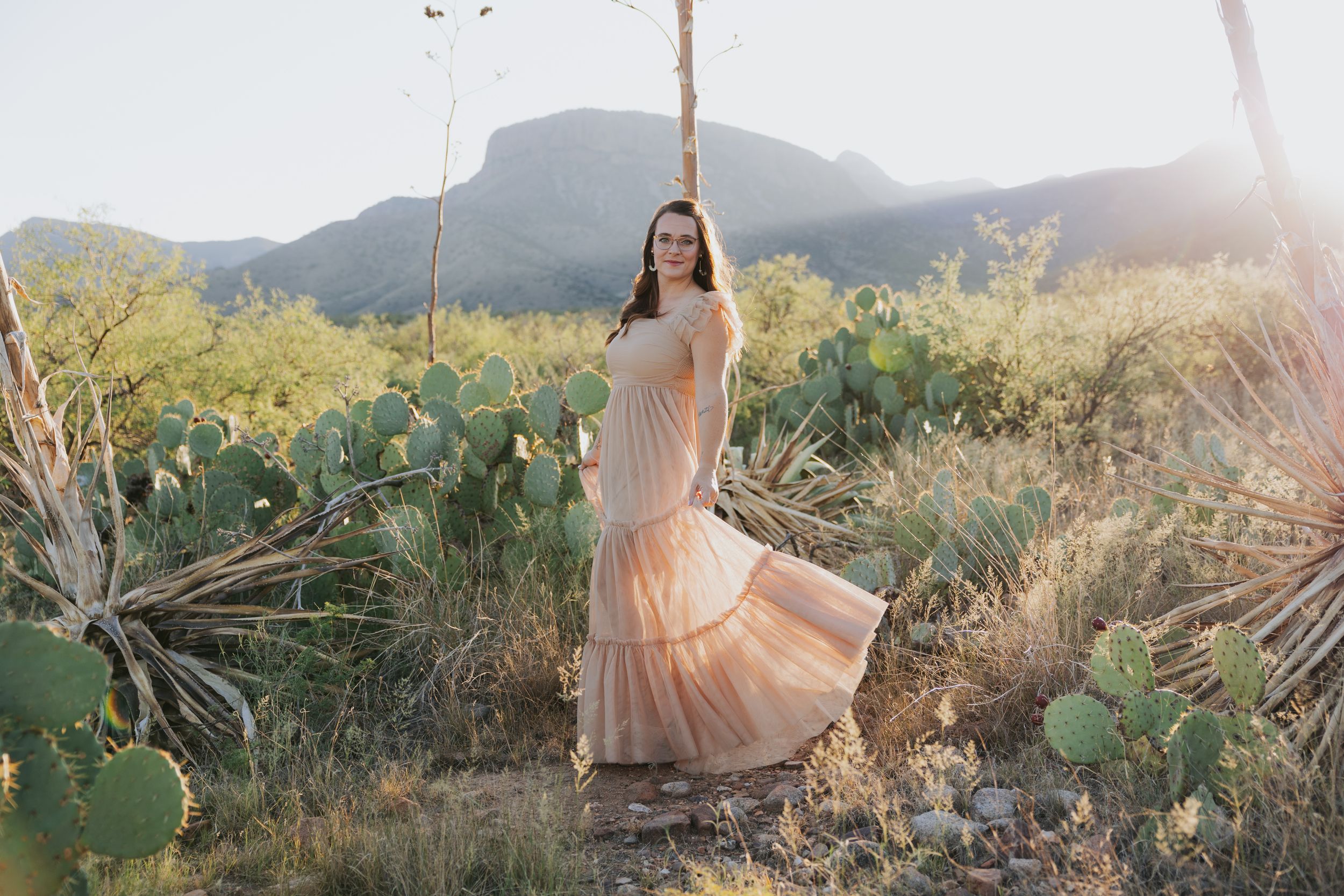 Flowing dress catches sunlight in desert landscape with mountains and cacti.