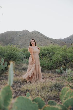 A romantic desert photoshoot features a flowing peach dress among cacti and mountains.
