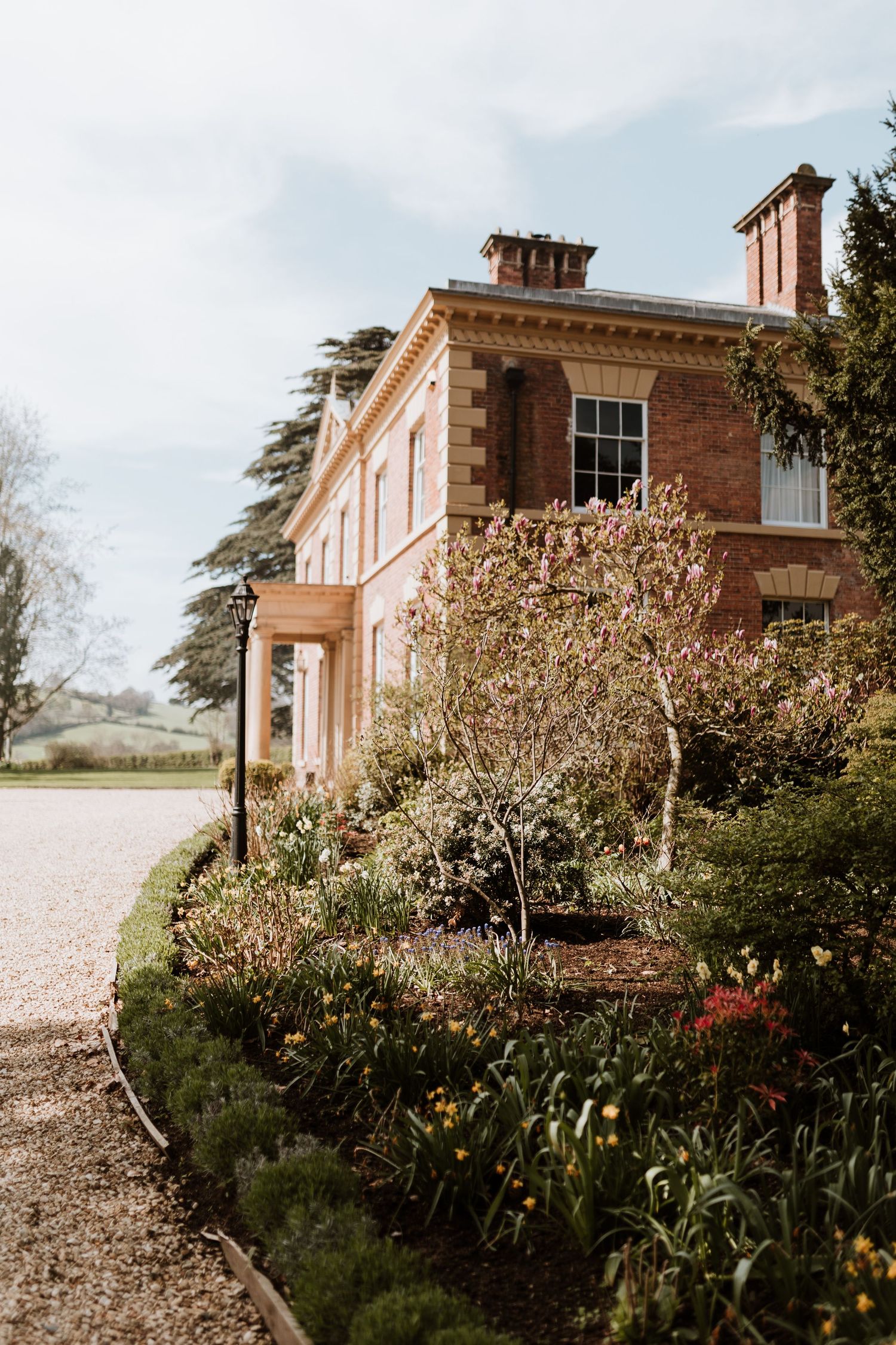 A rustic brick mansion house with blooming garden beds and spring tulips along the landscaped pathway.
