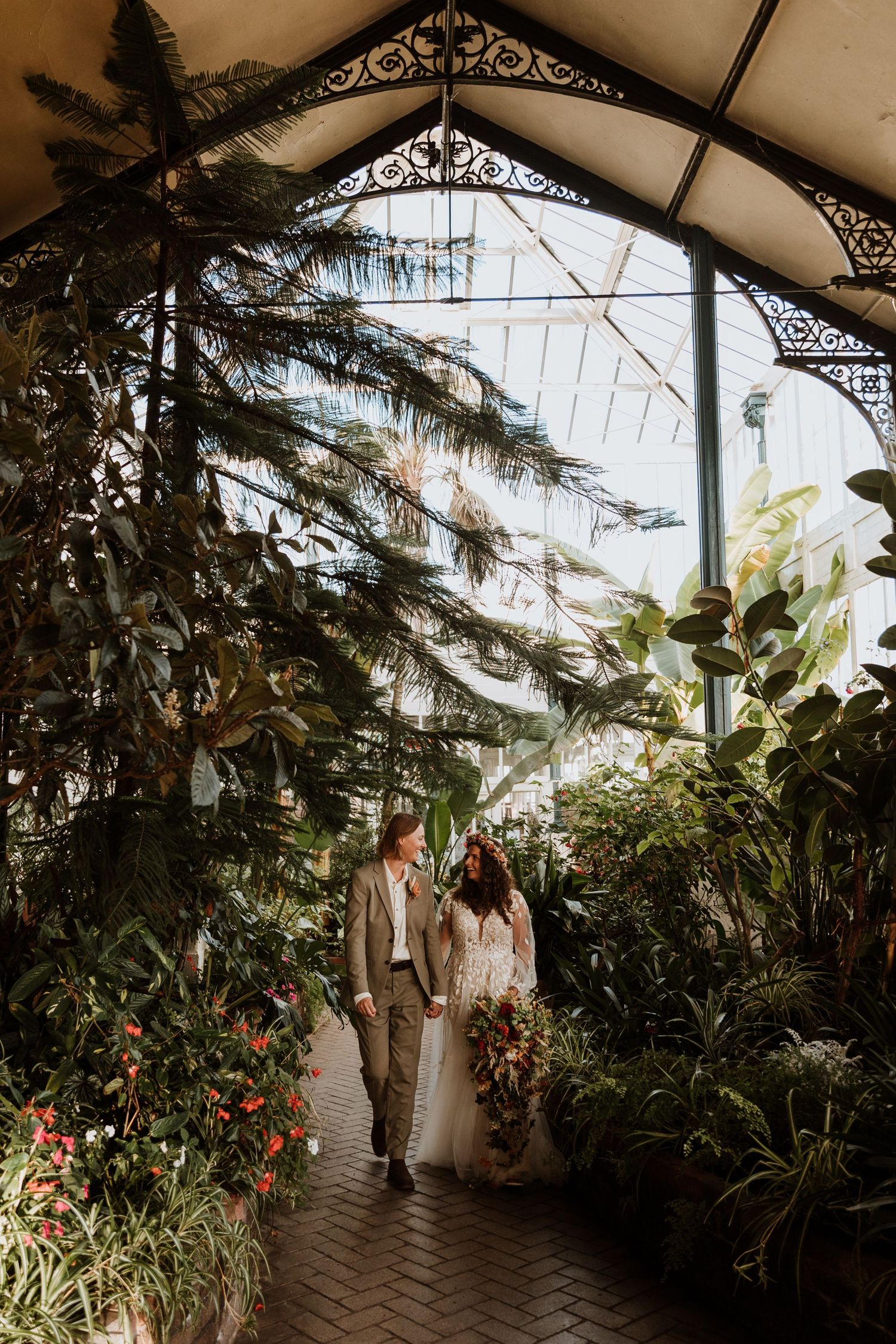 A lush greenhouse walkway with ornate ironwork ceiling, abundant tropical plants and natural lighting creating a botanical ambiance.