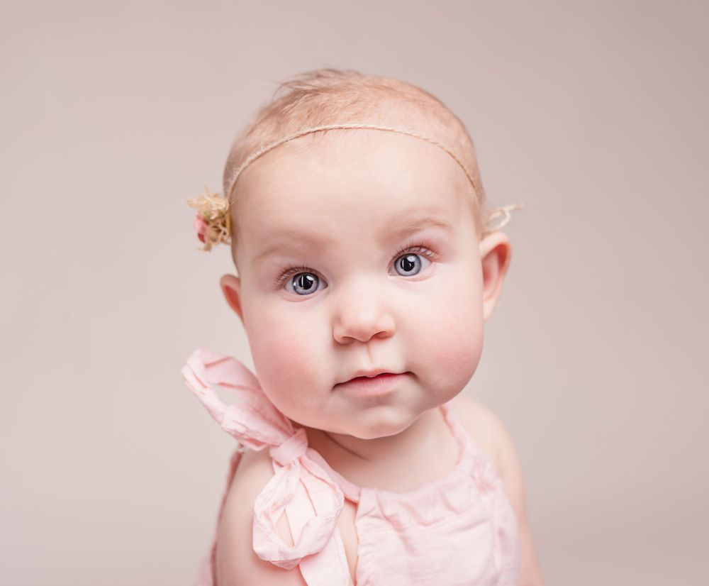 Baby wearing pink bow headband and dress poses for portrait against neutral background.